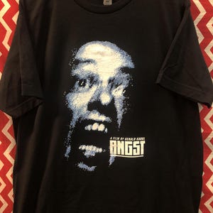 May include: Black t-shirt featuring a blue and white graphic of a face screaming. The text "A film by Gerald Kargl" and "ANGST" are printed below the face.