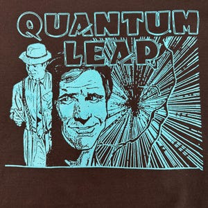 May include: A black t-shirt with a blue and black graphic print of a man in a fedora and a man's face. The text "QUANTUM LEAP" is printed in blue above the graphic.