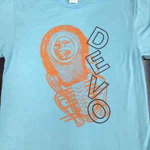 Op de afbeelding: Een lichtblauw t-shirt met een oranje en zwarte afbeelding van een robotkop en het woord "DEVO" gedrukt op de voorkant.