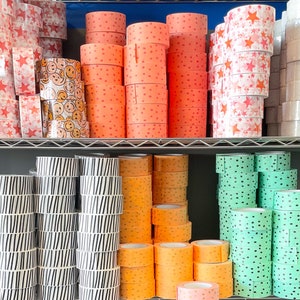 Puede incluir: Un estante lleno de rollos de cinta washi estampada de colores. La cinta viene en una variedad de colores y patrones, incluyendo lunares, rayas, estrellas y caritas sonrientes.