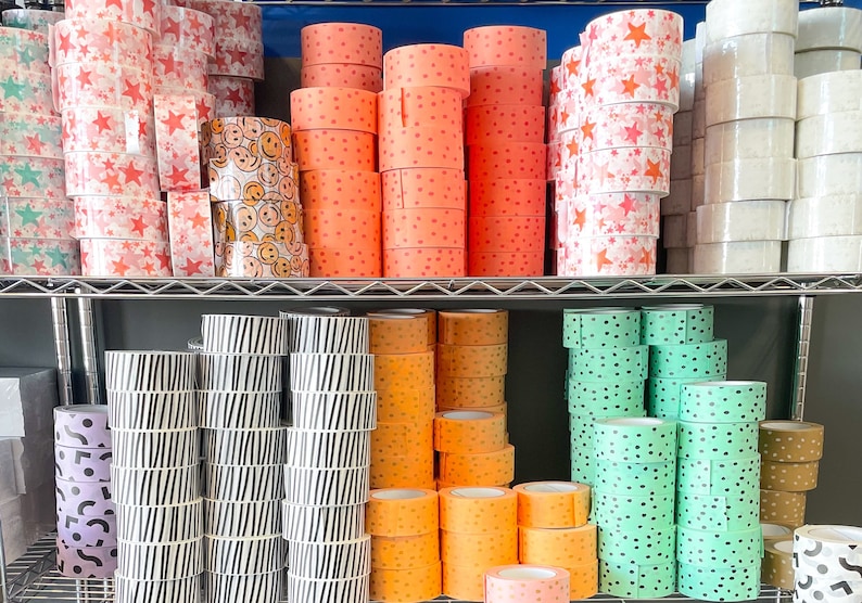 Puede incluir: Un estante lleno de rollos de cinta washi estampada de colores. La cinta viene en una variedad de colores y patrones, incluyendo lunares, rayas, estrellas y caritas sonrientes.