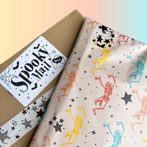 Puede incluir: Una caja de cart&oacute;n marr&oacute;n con una pegatina blanca que dice "Spooky Mail!" y un dise&ntilde;o de telara&ntilde;a en blanco y negro. La caja est&aacute; envuelta en papel de seda blanco con un patr&oacute;n repetido de esqueletos bailando y estrellas negras.