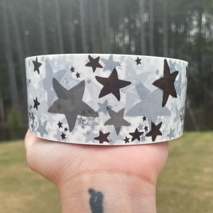 Puede incluir: Un rollo de cinta washi blanca con un patr&oacute;n de estrellas negras y grises. Las estrellas son de diferentes tama&ntilde;os y est&aacute;n dispersas por la cinta.