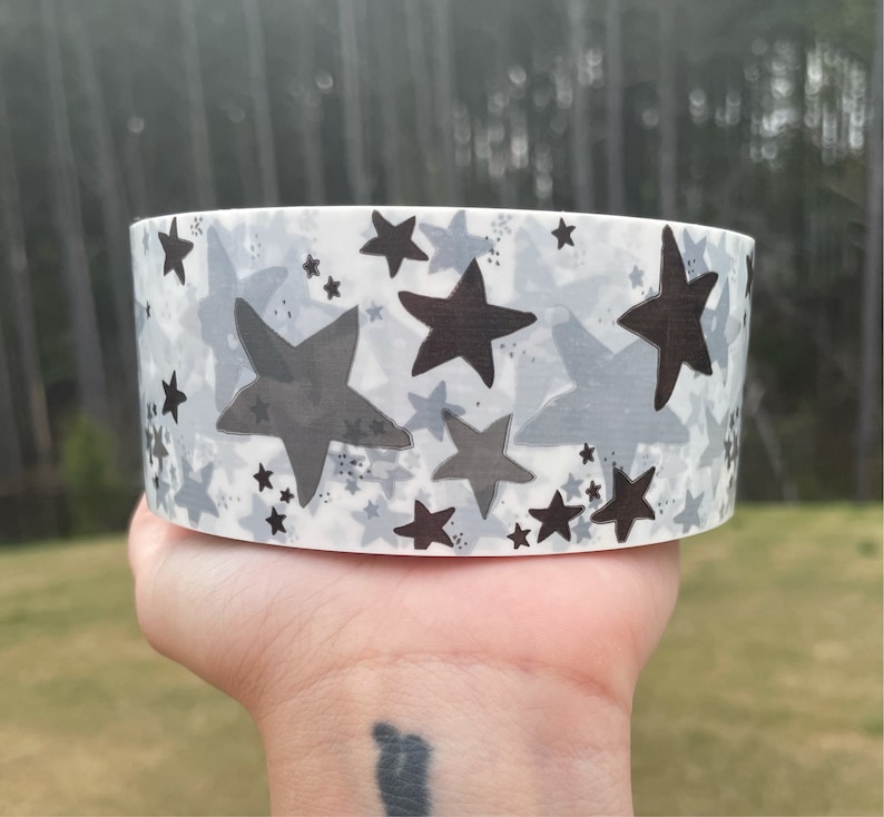 Puede incluir: Un rollo de cinta washi blanca con un patr&oacute;n de estrellas negras y grises. Las estrellas son de diferentes tama&ntilde;os y est&aacute;n dispersas por la cinta.