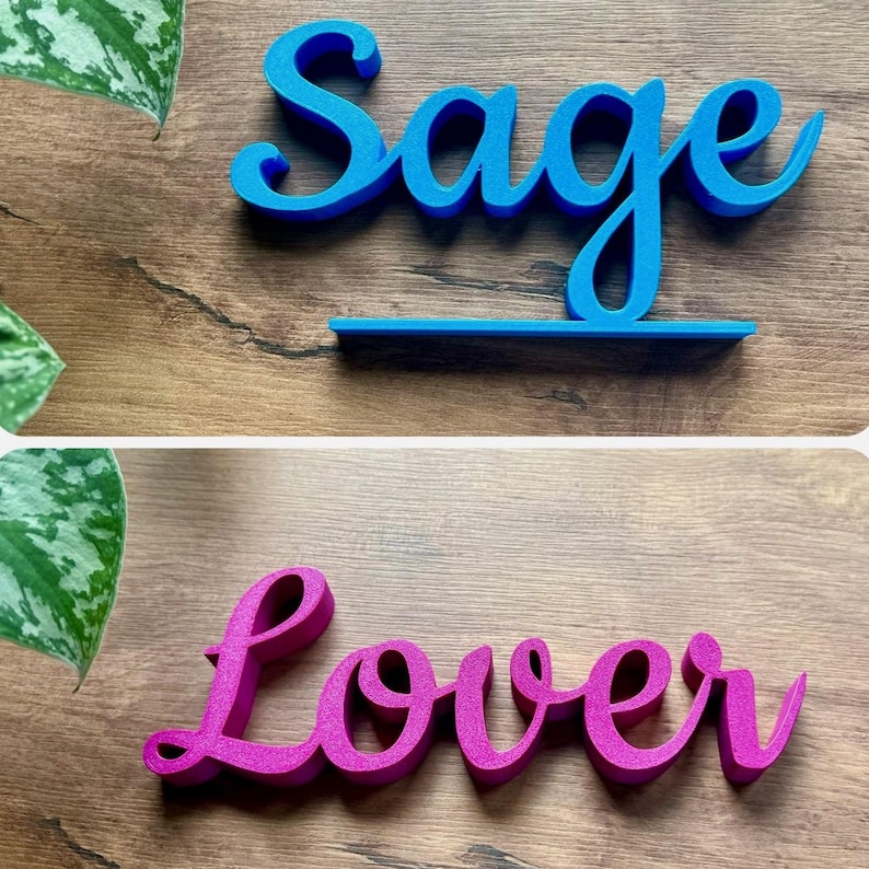 May include: Two wooden names, "Sage" in blue and "Lover" in pink, are displayed on a woodgrain surface. The names are in a cursive font and are 3D.