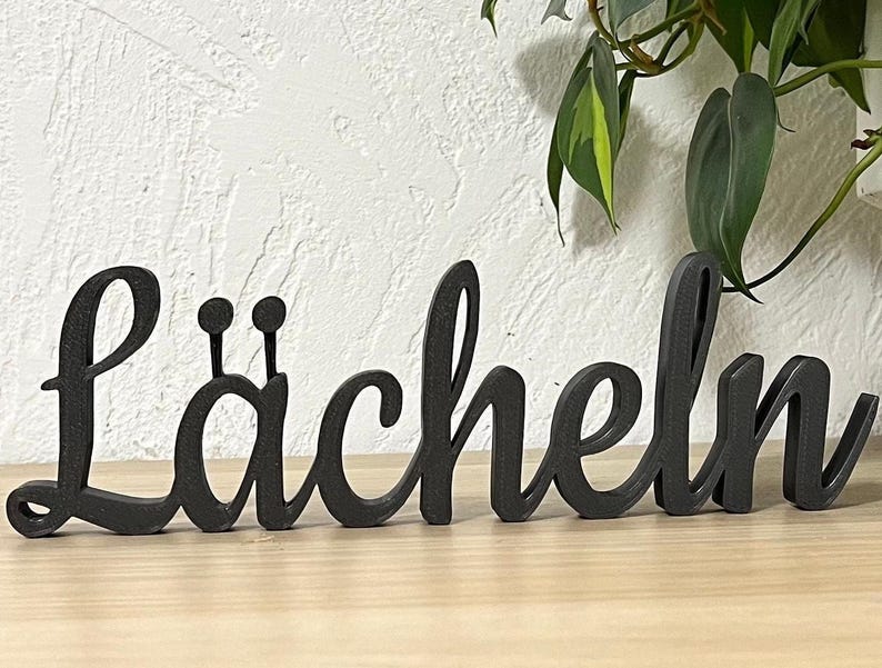 May include: Black wooden sign with the word "L&auml;cheln" in a cursive font.