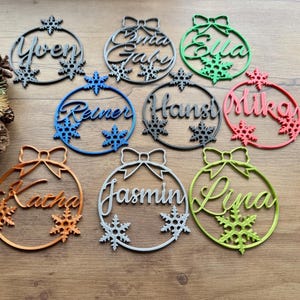 Puede incluir: Conjunto de etiquetas navideñas con forma de adorno, con nombres decorativos y detalles de copos de nieve. Los colores incluyen verde, rojo, azul, naranja y gris. Cada adorno tiene un lazo.