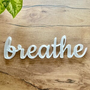 May include: White cursive word "breathe" on a wood background.