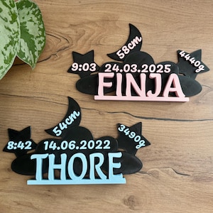 Op de afbeelding: Twee zwart-witte baby-aankondigingsplaques met een wolk- en sterrendesign. De bovenste plaquette is roze en zegt "FINJA" met de datum "24.03.2025", de tijd "9:03" en het gewicht "4440g". De onderste plaquette is blauw en zegt "THORE" met de datum "14.06.2022", de tijd "8:42" en het gewicht "3490g".
