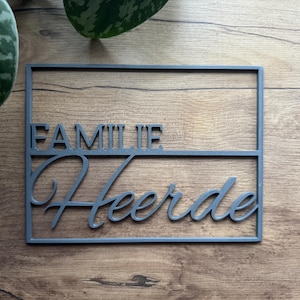 May include: A gray rectangular sign with the word "FAMILIE" above "Heerde" in a cursive font. The sign is made of a solid material and is set against a wooden background.