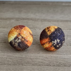 May include: A pair of fabric earrings with a colorful abstract pattern in shades of orange, yellow, and brown. The earrings are round and have a slightly textured surface.