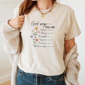 May include: A beige t-shirt with the text "God says YOU ARE" and inspirational words like "Unique" and "Strong" with floral accents. The shirt is paired with blue jeans and a beige cardigan.