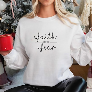 Può includere: Felpa bianca con la scritta "Faith over Fear" in corsivo nero. La felpa è a girocollo e sembra essere realizzata in un materiale morbido. L'immagine è scattata in interni, con un albero di Natale sullo sfondo.
