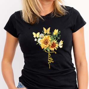Könnte beinhalten: Schwarzes T-Shirt mit einem Blumendesign aus Sonnenblumen, Schmetterlingen und Gänseblümchen in Gelb und Weiß. Das Wort "faith" ist in gelber Schrift geschrieben. Das T-Shirt hat kurze Ärmel.