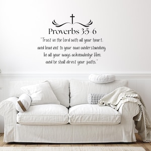 May include: A white sofa with a matching slipcover is positioned against a white wall. Above the sofa, black text reads "Proverbs 3:5-6" and a quote. A potted plant and a rug are in the foreground. Decorative pillows and a blanket adorn the sofa.
