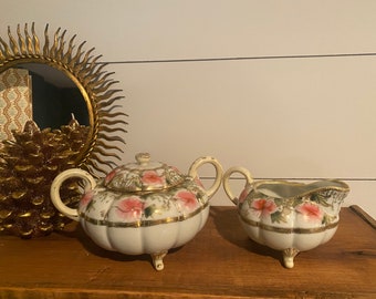 May include: A vintage porcelain sugar and creamer set with a floral design. The pieces are white with pink and gold accents. The sugar bowl has a lid and both pieces have three feet.