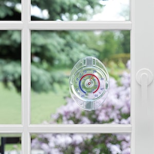 Może przedstawiać: Okrągły termometr z przezroczystego plastiku z białą tarczą i czerwonymi i niebieskimi oznaczeniami. Termometr jest przymocowany do okna za pomocą przyssawki.