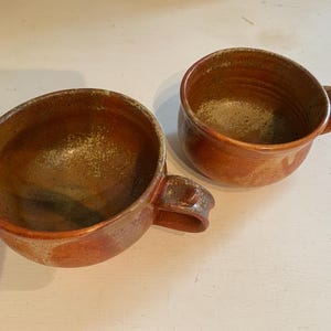 May include: Two brown ceramic mugs with a speckled glaze. The mugs have a rounded shape and a handle.