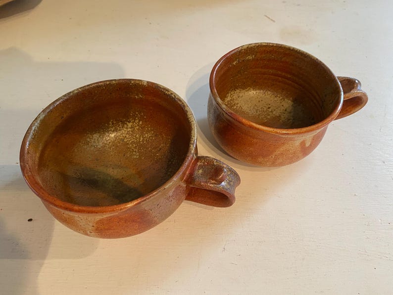 May include: Two brown ceramic mugs with a speckled glaze. The mugs have a rounded shape and a handle.