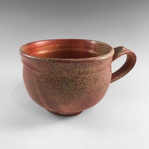 May include: A handmade ceramic mug with a rounded body and a single handle. The mug has a speckled brown and red glaze.