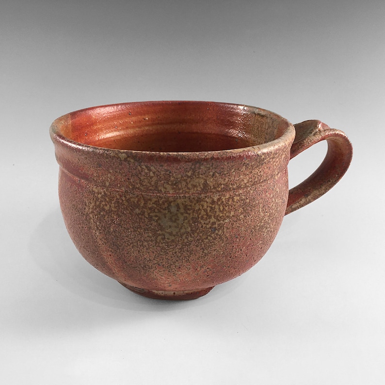 May include: A handmade ceramic mug with a rounded body and a single handle. The mug has a speckled brown and red glaze.