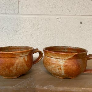 May include: Two brown ceramic mugs with a speckled glaze and a rounded shape. The mugs have a handle on one side.