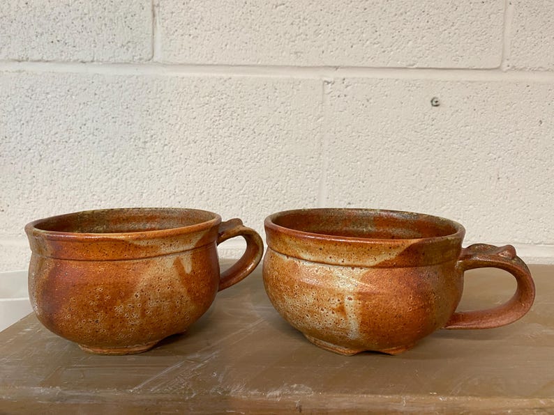 May include: Two brown ceramic mugs with a speckled glaze and a rounded shape. The mugs have a handle on one side.