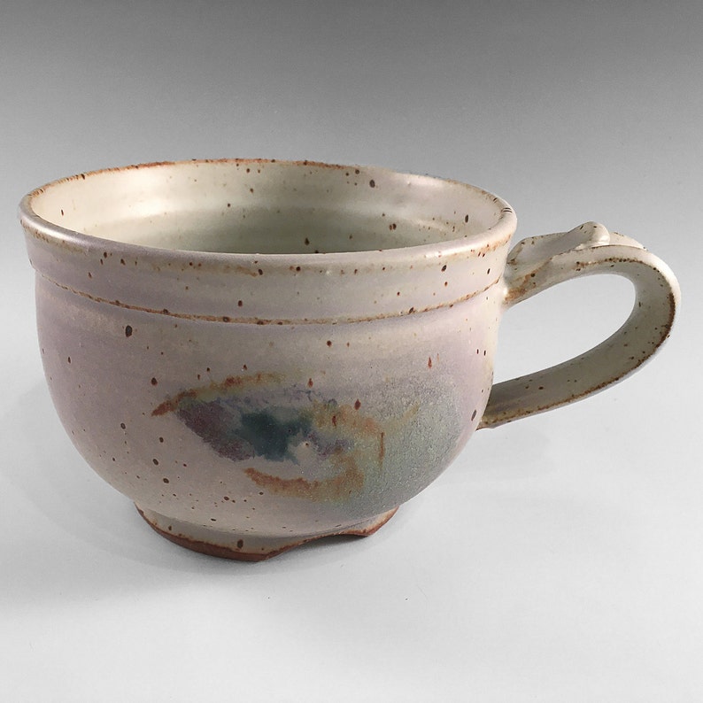 May include: A white ceramic mug with a speckled glaze and a brown, green, and purple design. The mug has a rounded shape and a handle.
