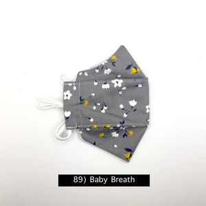 Może przedstawiać: Szara maska ​​na twarz z białym i ż&oacute;łtym wzorem kwiatowym. Maska ​​ma białe sznurki. Tekst "89) Baby Breath" jest nadrukowany pod maską.