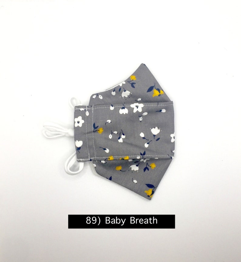 Może przedstawiać: Szara maska ​​na twarz z białym i ż&oacute;łtym wzorem kwiatowym. Maska ​​ma białe sznurki. Tekst "89) Baby Breath" jest nadrukowany pod maską.