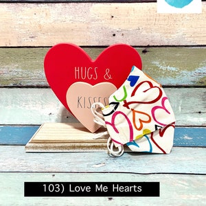 Puede incluir: Un coraz&oacute;n de madera rojo con el texto "Hugs & Kisses" en &eacute;l, y una mascarilla de tela blanca con corazones y remolinos coloridos. La mascarilla est&aacute; sobre una superficie de madera. El texto "103) Love Me Hearts" est&aacute; debajo de la imagen.