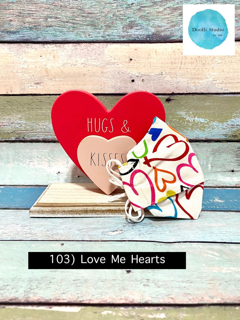 Puede incluir: Un coraz&oacute;n de madera rojo con el texto "Hugs & Kisses" en &eacute;l, y una mascarilla de tela blanca con corazones y remolinos coloridos. La mascarilla est&aacute; sobre una superficie de madera. El texto "103) Love Me Hearts" est&aacute; debajo de la imagen.