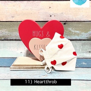 Puede incluir: Una mascarilla facial blanca con corazones rojos impresos en ella. La mascarilla est&aacute; sentada sobre un letrero de madera en forma de coraz&oacute;n que dice "Hugs & Kisses".