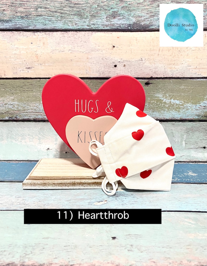 Puede incluir: Una mascarilla facial blanca con corazones rojos impresos en ella. La mascarilla est&aacute; sentada sobre un letrero de madera en forma de coraz&oacute;n que dice "Hugs & Kisses".