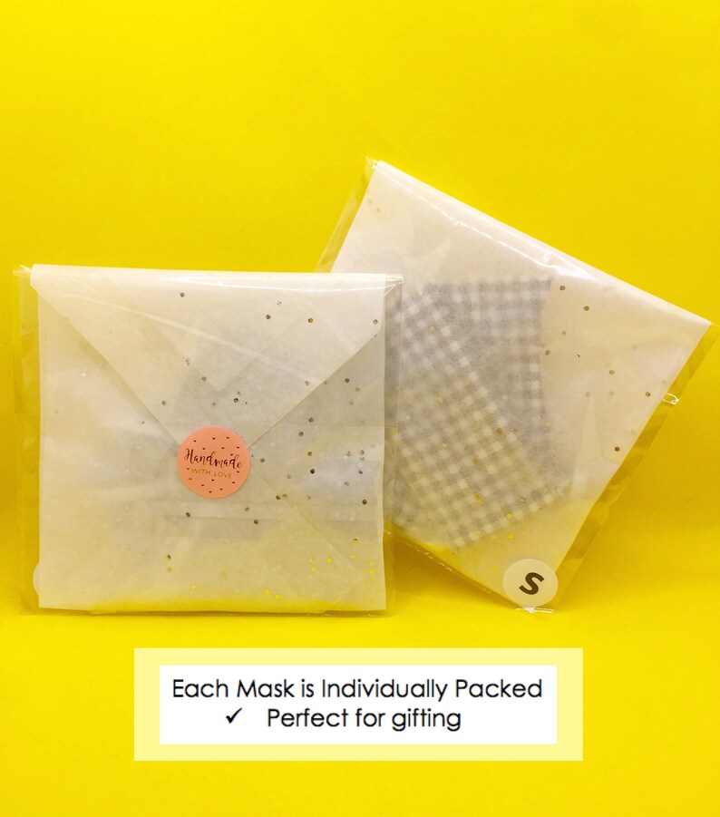 Puede incluir: Dos sobres de papel de seda blanco con puntos de purpurina dorada, uno con una pegatina rosa "Handmade with Love" y el otro con una tela de cuadros blancos y grises en el interior. El texto "Each Mask is Individually Packed" y una marca de verificaci&oacute;n con el texto "Perfect for gifting" est&aacute;n impresos sobre un fondo amarillo.