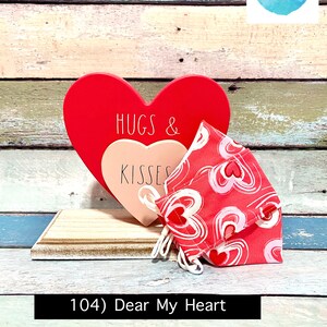 Puede incluir: Un coraz&oacute;n de madera rojo con el texto "Hugs & Kisses" en &eacute;l, y una mascarilla de tela rosa y roja con un patr&oacute;n de coraz&oacute;n. El coraz&oacute;n y la mascarilla est&aacute;n sentados en un estante de madera. El texto "104) Dear My Heart" est&aacute; escrito debajo del estante.