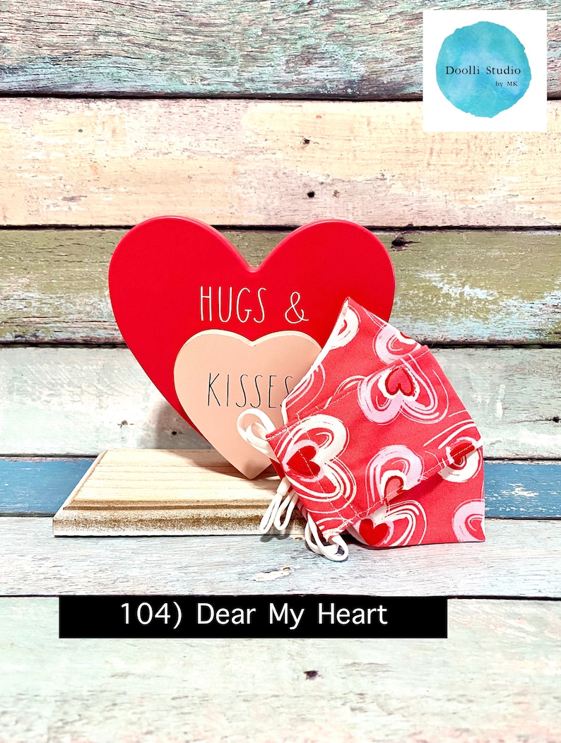 Puede incluir: Un coraz&oacute;n de madera rojo con el texto "Hugs & Kisses" en &eacute;l, y una mascarilla de tela rosa y roja con un patr&oacute;n de coraz&oacute;n. El coraz&oacute;n y la mascarilla est&aacute;n sentados en un estante de madera. El texto "104) Dear My Heart" est&aacute; escrito debajo del estante.