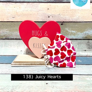 Puede incluir: Un coraz&oacute;n de madera rojo y rosa con el texto "Hugs & Kisses" en &eacute;l. Una mascarilla facial blanca con corazones rosas y rojos est&aacute; sentada sobre una superficie de madera. El texto "138) Juicy Hearts" est&aacute; debajo de la mascarilla.