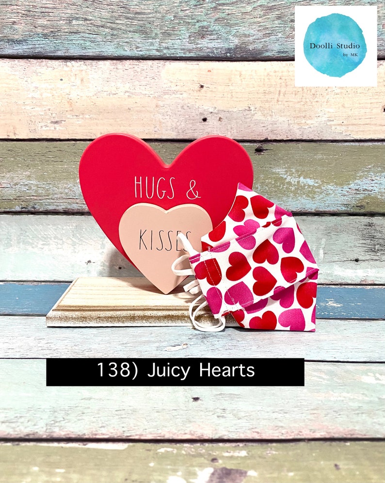 Puede incluir: Un coraz&oacute;n de madera rojo y rosa con el texto "Hugs & Kisses" en &eacute;l. Una mascarilla facial blanca con corazones rosas y rojos est&aacute; sentada sobre una superficie de madera. El texto "138) Juicy Hearts" est&aacute; debajo de la mascarilla.