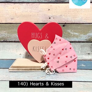 Puede incluir: Un letrero de madera en forma de coraz&oacute;n rosa y rojo con el texto "HUGS & KISSES" en &eacute;l. Una mascarilla facial rosa con un patr&oacute;n de peque&ntilde;os corazones y cruces est&aacute; delante del letrero. El letrero y la mascarilla est&aacute;n sobre una superficie de madera.