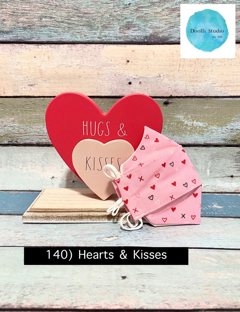 Puede incluir: Un letrero de madera en forma de coraz&oacute;n rosa y rojo con el texto "HUGS & KISSES" en &eacute;l. Una mascarilla facial rosa con un patr&oacute;n de peque&ntilde;os corazones y cruces est&aacute; delante del letrero. El letrero y la mascarilla est&aacute;n sobre una superficie de madera.
