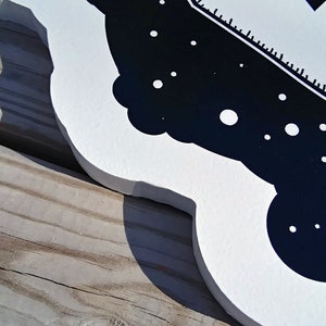 May include: A white wooden sign with a black and white design. The design features a stylized image of a ship, with a dark blue background and white accents. The sign is placed on a wooden surface.