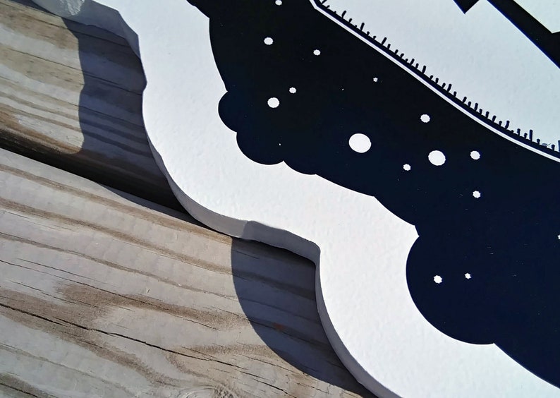 May include: A white wooden sign with a black and white design. The design features a stylized image of a ship, with a dark blue background and white accents. The sign is placed on a wooden surface.