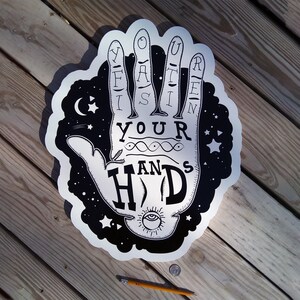 May include: A black and white graphic of a hand with the words "Lift Your Hands" and "Hold" in a stylized font. The design includes stars, a crescent moon, and an eye symbol. The artwork is on a silver background.