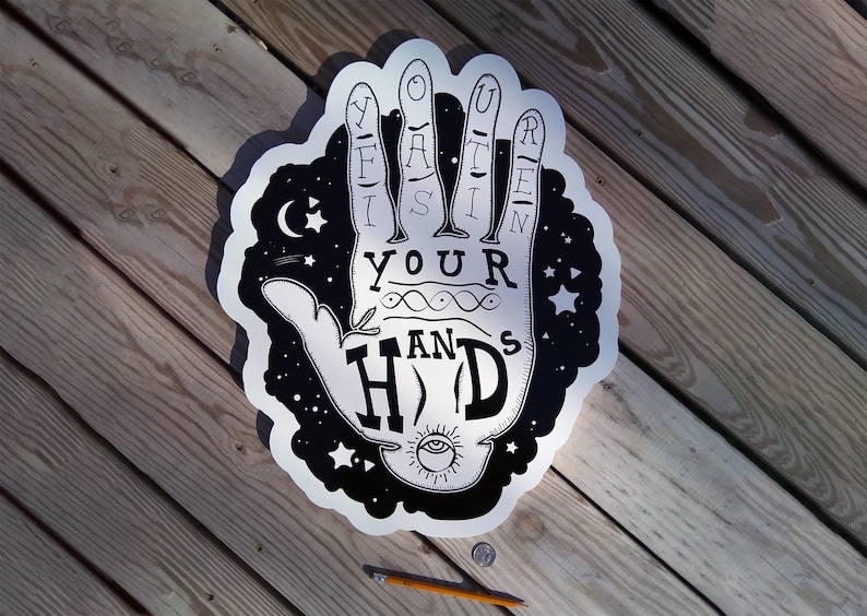 May include: A black and white graphic of a hand with the words "Lift Your Hands" and "Hold" in a stylized font. The design includes stars, a crescent moon, and an eye symbol. The artwork is on a silver background.