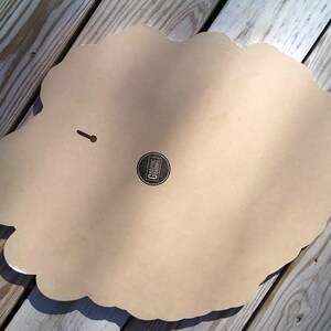 May include: A decorative wall hanging with a scalloped edge, viewed from the back. The piece is a light tan color with a small circular logo and a keyhole for hanging. The surface is smooth and the edges are white.