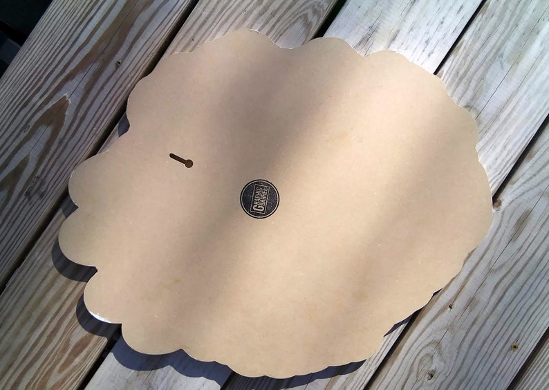 May include: A decorative wall hanging with a scalloped edge, viewed from the back. The piece is a light tan color with a small circular logo and a keyhole for hanging. The surface is smooth and the edges are white.