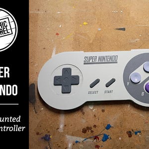 May include: A wall-mounted Super Nintendo game controller. The controller is light gray with a directional pad, select and start buttons, and four colored buttons. The text "SUPER NINTENDO" is printed on the controller.