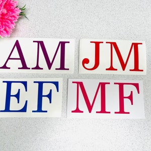 May include: Four rectangular decals with large, bold letters in various colours. The letters are AM in purple, JM in red, EF in blue, and MF in pink. A pink and white flower arrangement is in the upper left corner.