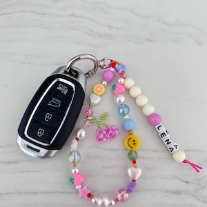 May include: A colorful beaded keychain with a smiley face charm, fruit charms, and a name tag that reads "LENA". The keychain is attached to a car key.